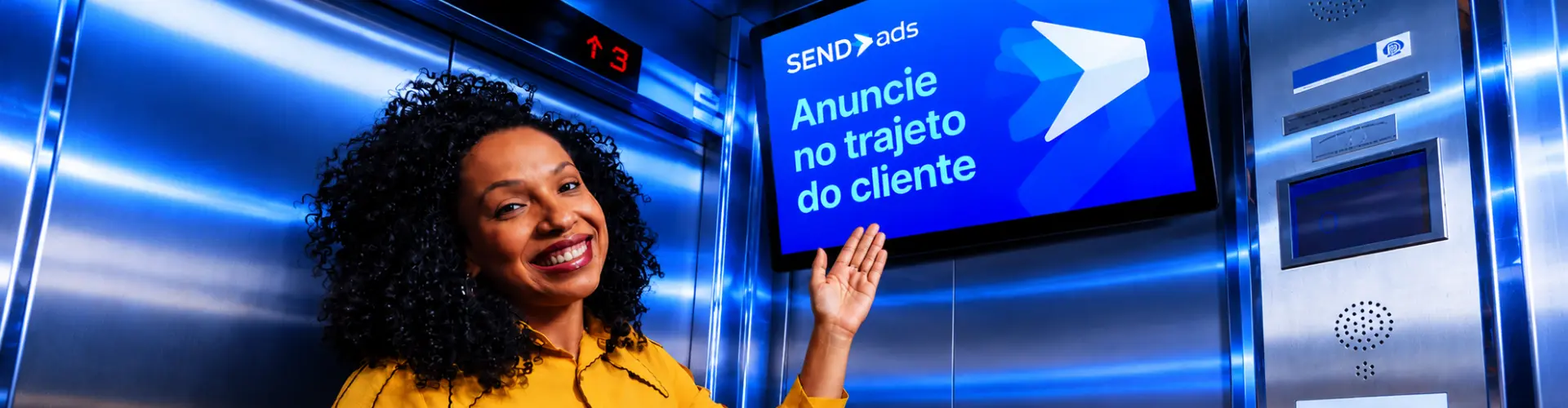 Mulher sorrindo dentro de um elevador aponta para a tela da SendADS com a frase: Anuncie no trajeto do cliente