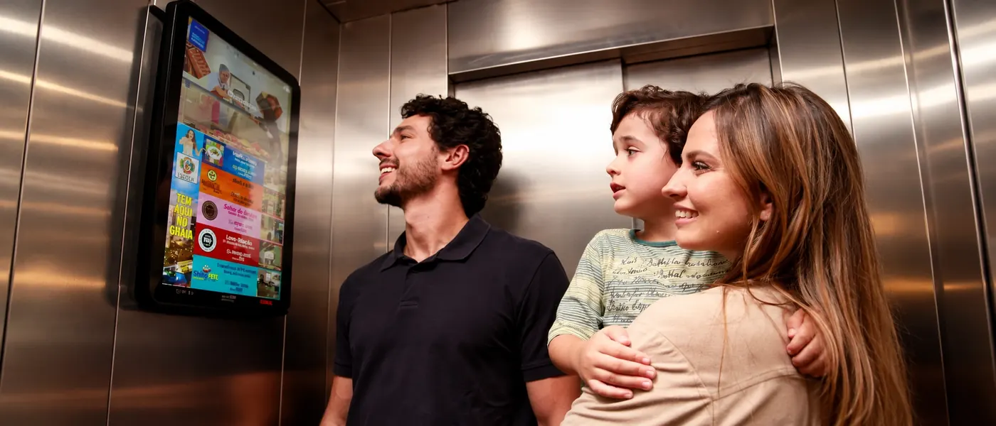 Família dentro do elevador olhando a tela digital com anúncios